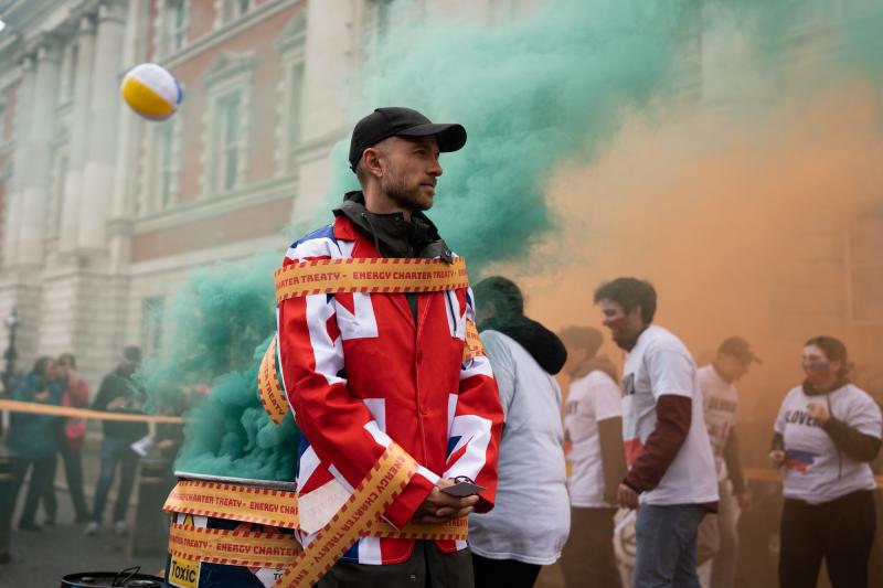 Person wearing a Union Jack jacket to symbolise the UK, tied to an oil barrel belching green smoke, with tape on which 'Energy Charter Treaty' is written